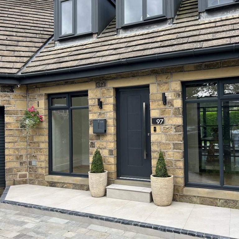 Moderner Eingangsbereich eines Hauses mit Steinfassade, Pflanzen und Glasfront.